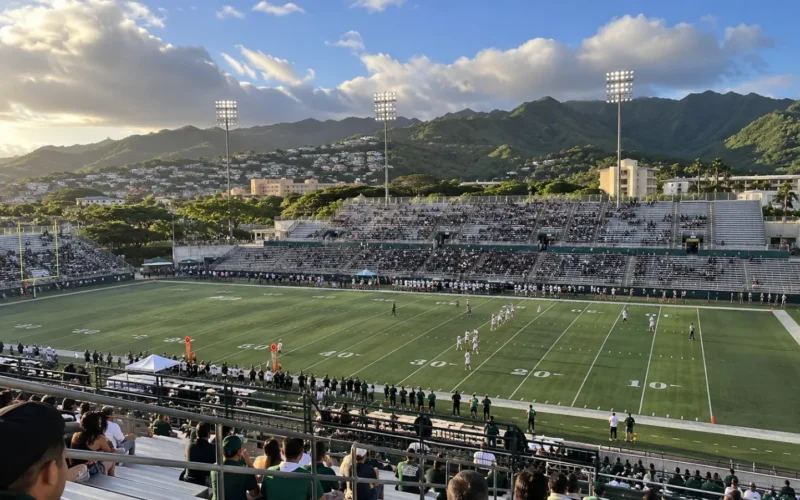 Football game at an open-air stadium below green Mānoa ridges at sunset