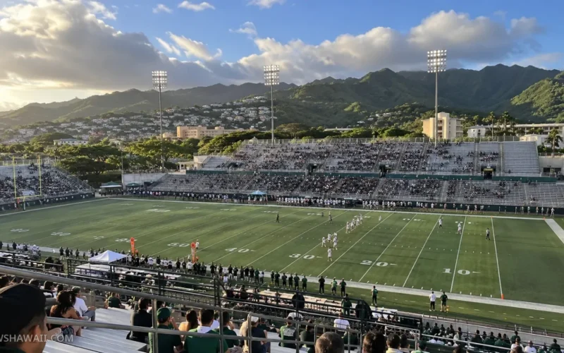 Football game at an open-air stadium below green Mānoa ridges at sunset