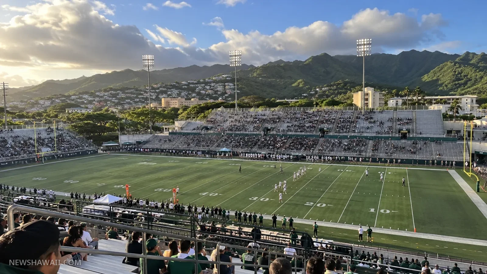 Football game at an open-air stadium below green Mānoa ridges at sunset